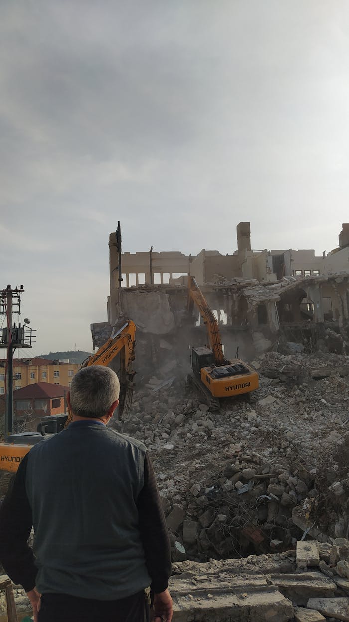 A man observes a building demolition with excavators in an urban setting.